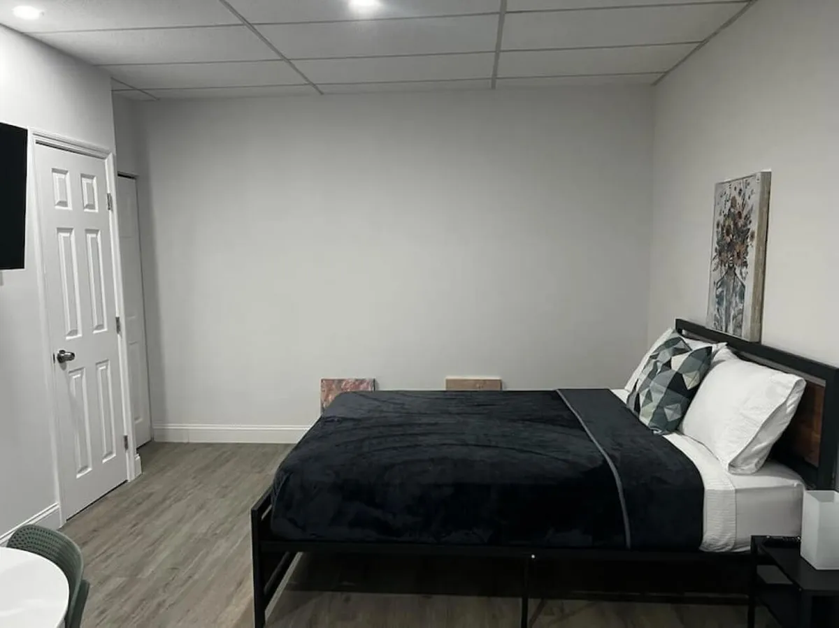 This image showcases a plush queen bed adorned with a soft blanket and decorative pillows, complemented by tasteful wall art above the headboard. A wall-mounted Smart TV is visible, and the room is bathed in natural light, creating an inviting and tranquil atmosphere perfect for both relaxation and productivity.