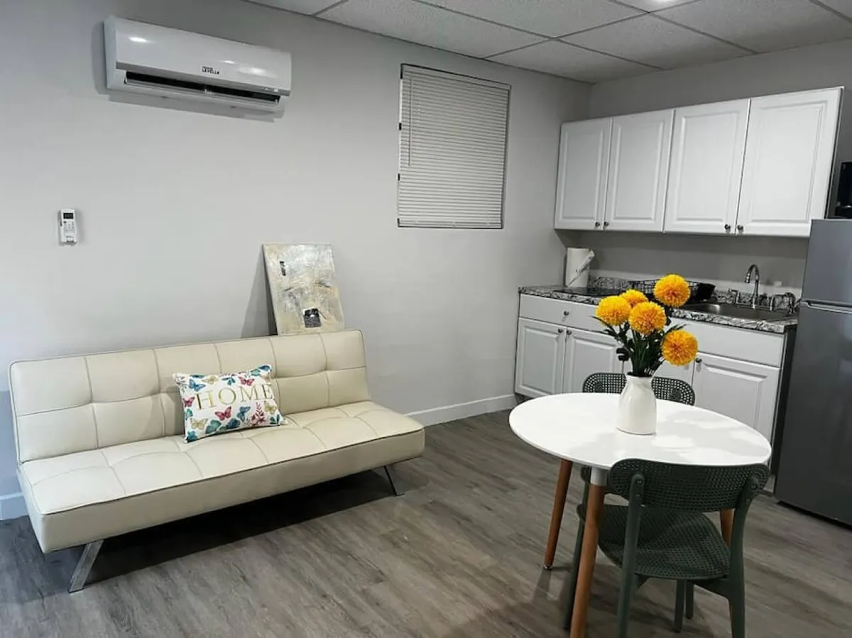 The image showcases a bright, inviting studio interior with a plush white leather sofa adorned with a 'HOME' pillow, a cozy round dining table set with cheerful yellow flowers, a compact kitchenette featuring modern white cabinetry and granite countertops, and a sleek stainless steel refrigerator. The room exudes a sense of warmth, comfort, and modern practicality, perfect for urban explorers.