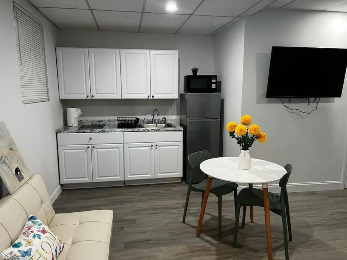 A bright, neatly organized studio featuring a cream-colored sofa with a decorative pillow, a small round dining table adorned with a vase of yellow flowers, white kitchen cabinetry with a sleek granite countertop, and a wall-mounted flat-screen TV. The space feels cozy and efficient, showcasing a blend of living, dining, and entertainment areas within the open-plan layout.